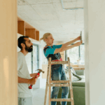 A man and a woman with a ladder and a hammer working on a home improvement project.