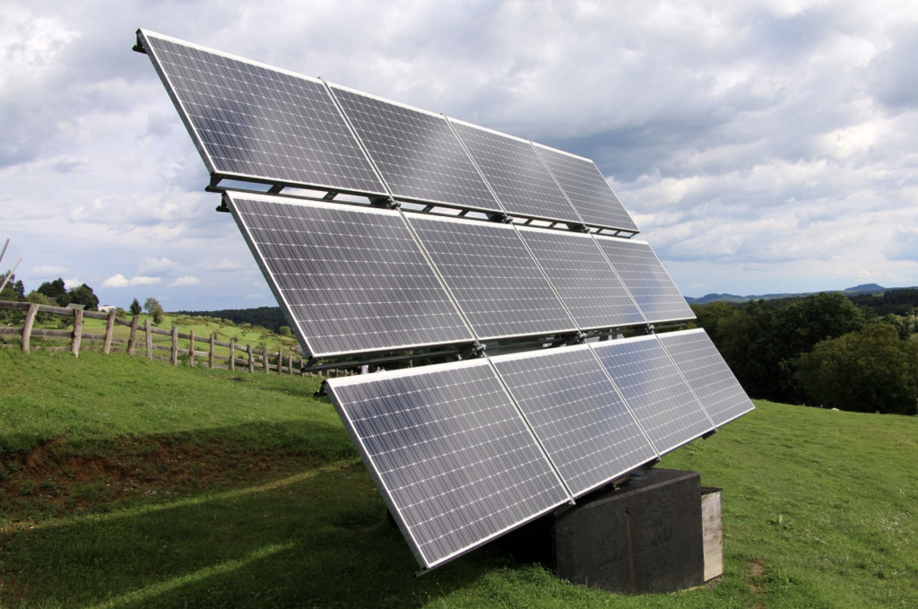 An array of solar panels placed at an angle, sitting on a grassy hill with fencing and trees in the background