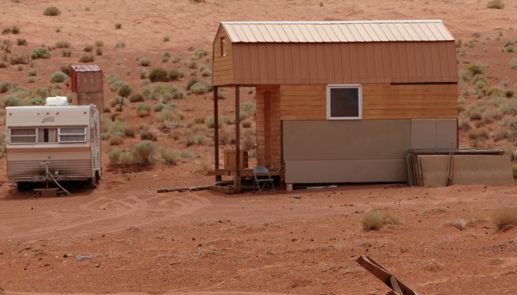 A prefabriacated tiny home sits on a red desert landscape with a travel trailer sitting to the left of it
