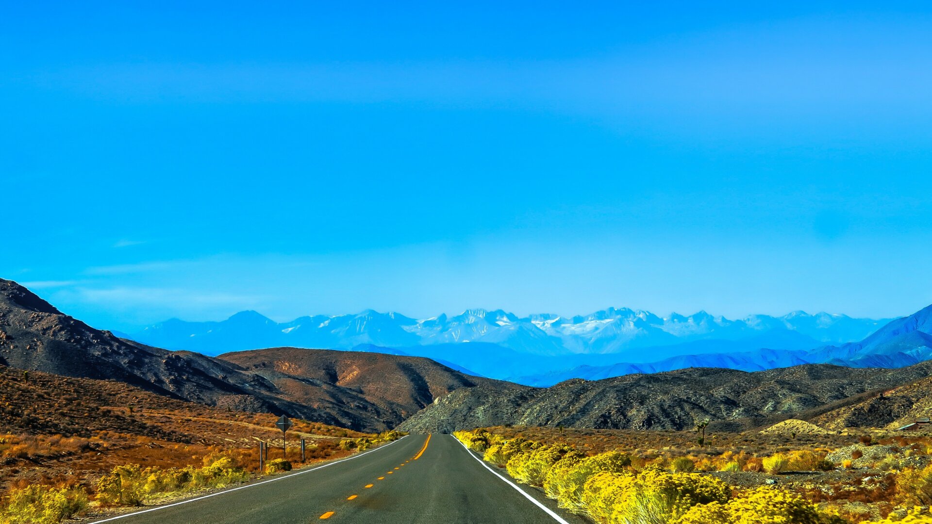 A desert mountain terrain with a road leading towards snow-capped mountains in the distance.