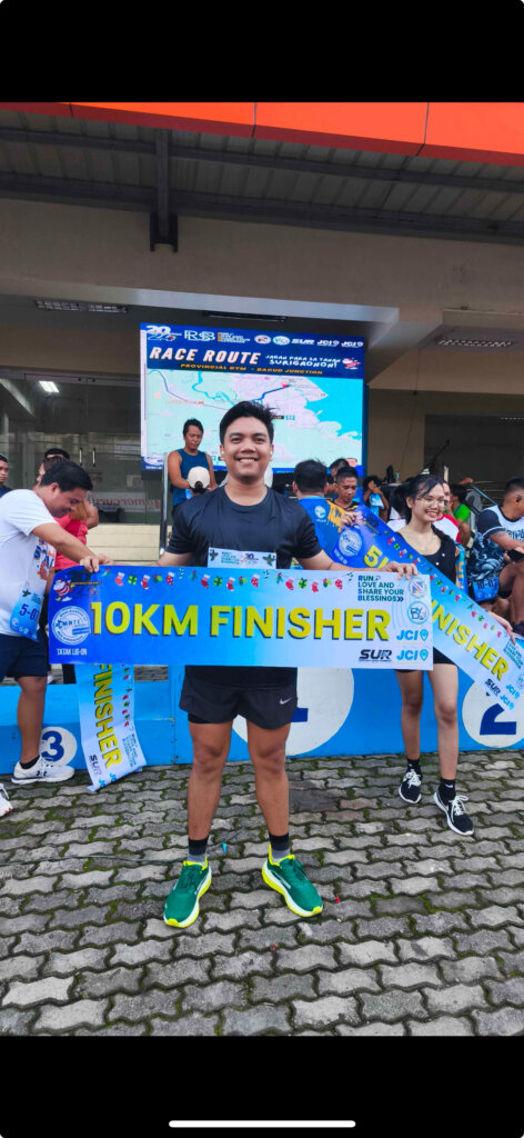John Pijoto smiling at the finish line of a 10K charity run, holding a 10KM Finisher banner