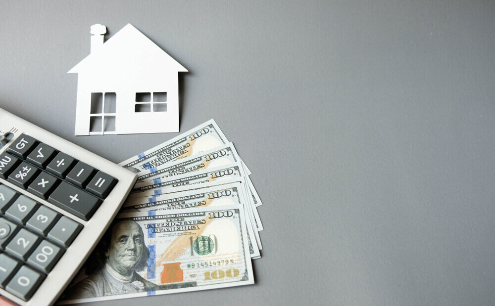 White paper house with dollars banknotes and calculator on a grey background.
