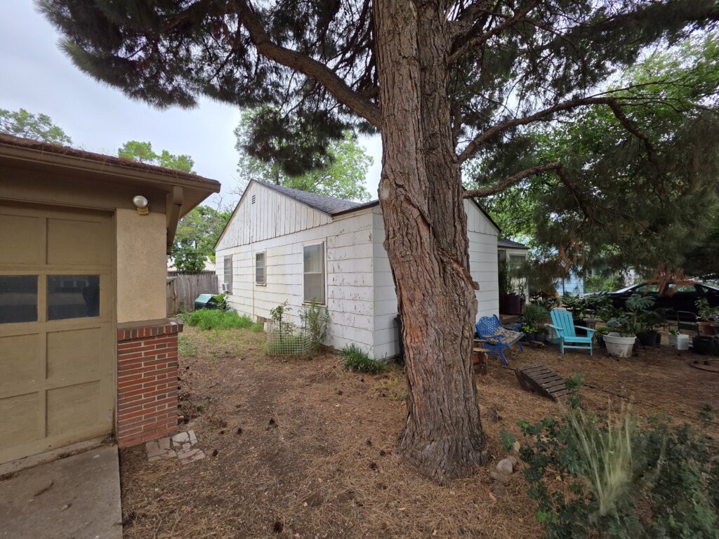 Older White House In Lubbock Texas With Peeling Paint, Large Tree In The Yard, And Outdoor Chairs, Showing That Cash Home Buyers Purchase Properties As-Is And Help Homeowners Sell Your House Fast Without Repairs Or Cleanup