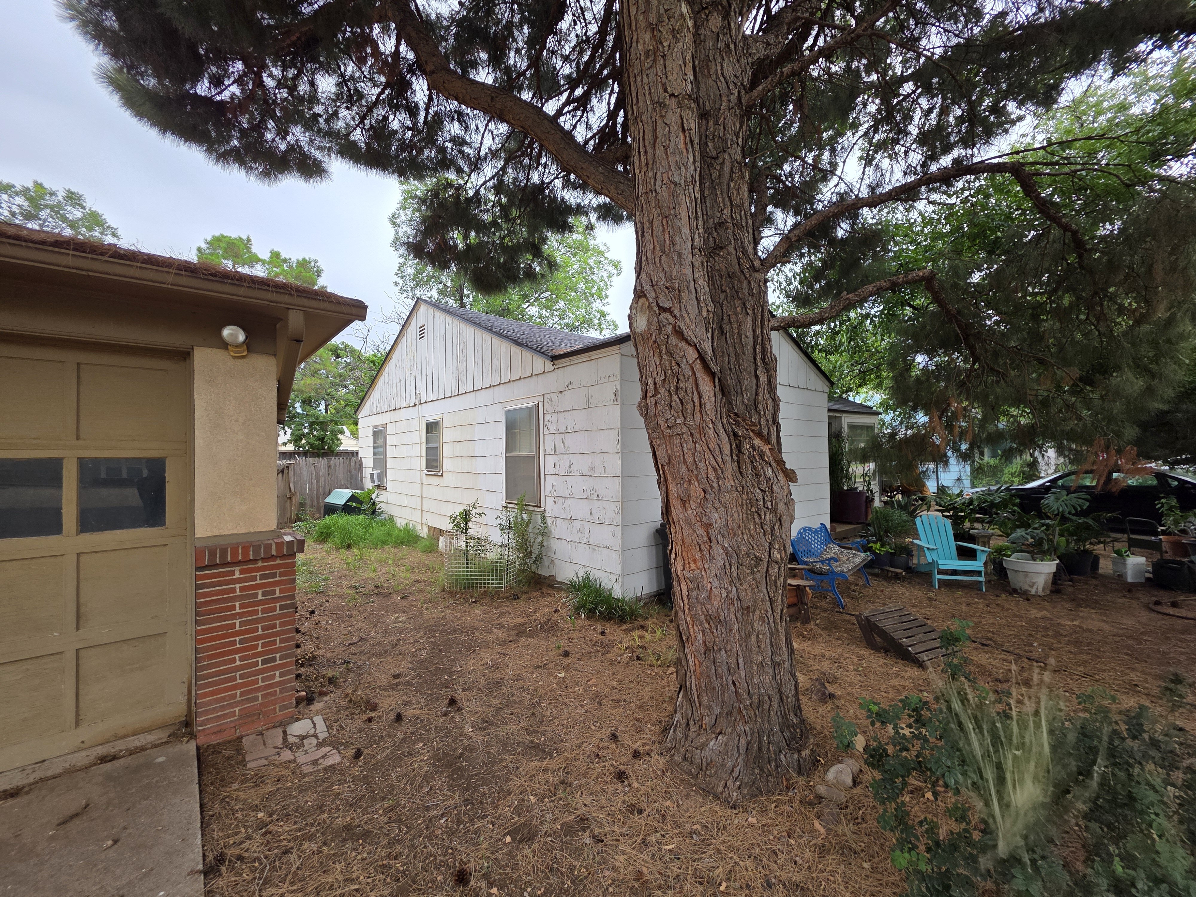 Older White House In Lubbock Texas With Peeling Paint, Large Tree In The Yard, And Outdoor Chairs, Showing That Cash Home Buyers Purchase Properties As-Is And Help Homeowners Sell Your House Fast Without Repairs Or Cleanup
