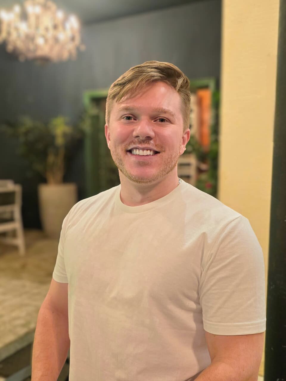 A smiling man wearing a light-colored T-shirt stands indoors under warm lighting, with a chandelier and soft background décor creating an inviting, professional atmosphere—used as a portrait for a real estate business owner.