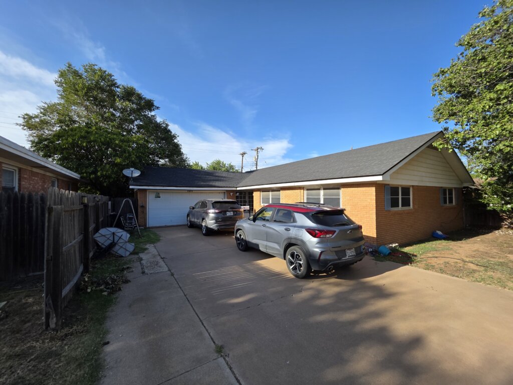 A brick single-story house in Lubbock, Texas with a two-car driveway leading to a white garage. Two vehicles are parked along the driveway, and the yard features mature trees and a wooden fence on the left side. The sky is bright and clear, creating a well-lit exterior view of the property.