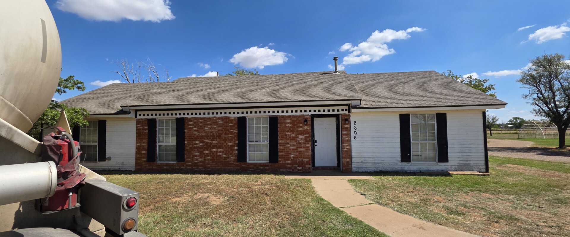 A single-story brick and white-siding house with black shutters under a bright blue sky in Lubbock, Texas. A curved concrete walkway leads to the front door, and a large utility truck is partially visible on the left side of the frame. The yard is dry with patches of grass, and open land with trees sits in the background.