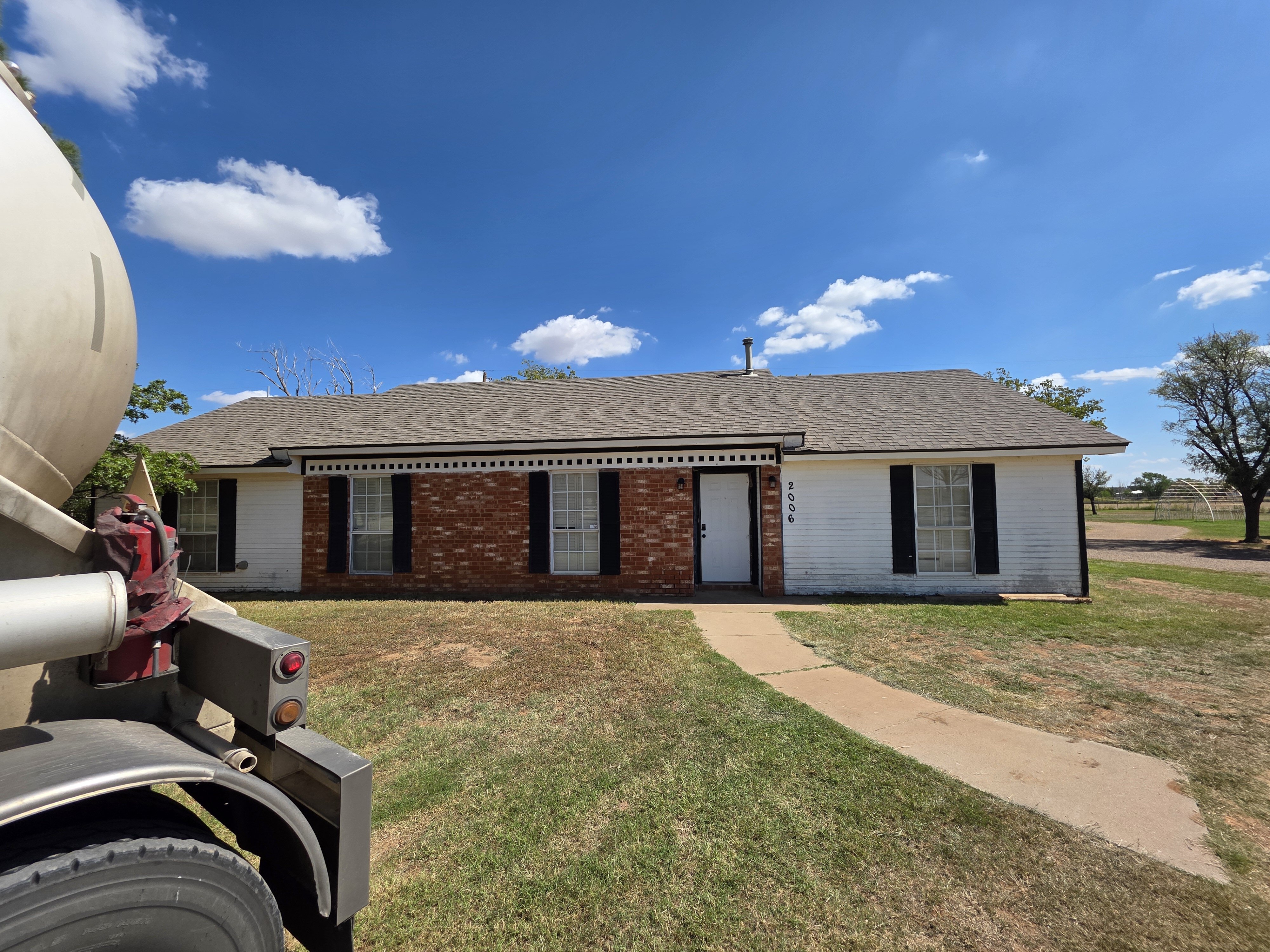 A single-story brick and white-siding house with black shutters under a bright blue sky in Lubbock, Texas. A curved concrete walkway leads to the front door, and a large utility truck is partially visible on the left side of the frame. The yard is dry with patches of grass, and open land with trees sits in the background.