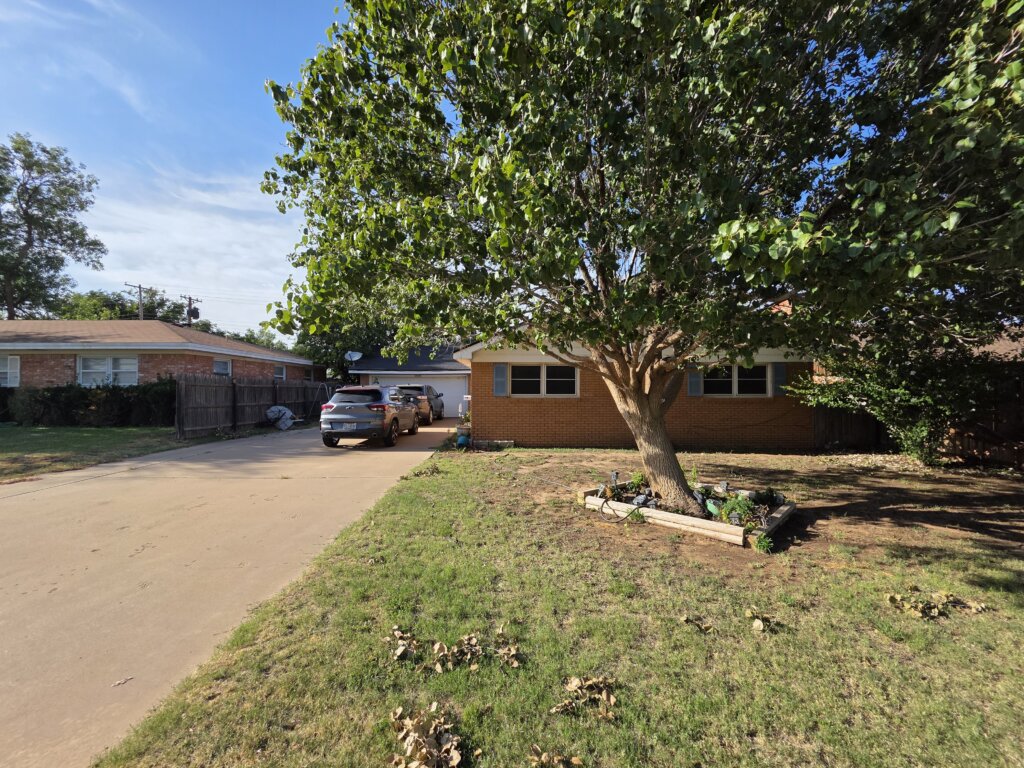 A single-story brick house in Lubbock, Texas with a large tree in the front yard casting shade over the lawn. Several vehicles are parked along the long driveway leading to the garage, and neighboring homes and fences are visible on each side under a bright blue sky.
