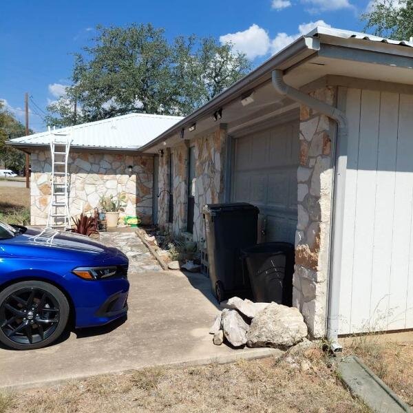 Side view of a stone house with a metal roof and an attached garage, showing an aging driveway, parked car, trash bins, and scattered rocks—highlighting a property that may need repairs and is ideal for a fast, as-is cash sale in Texas.