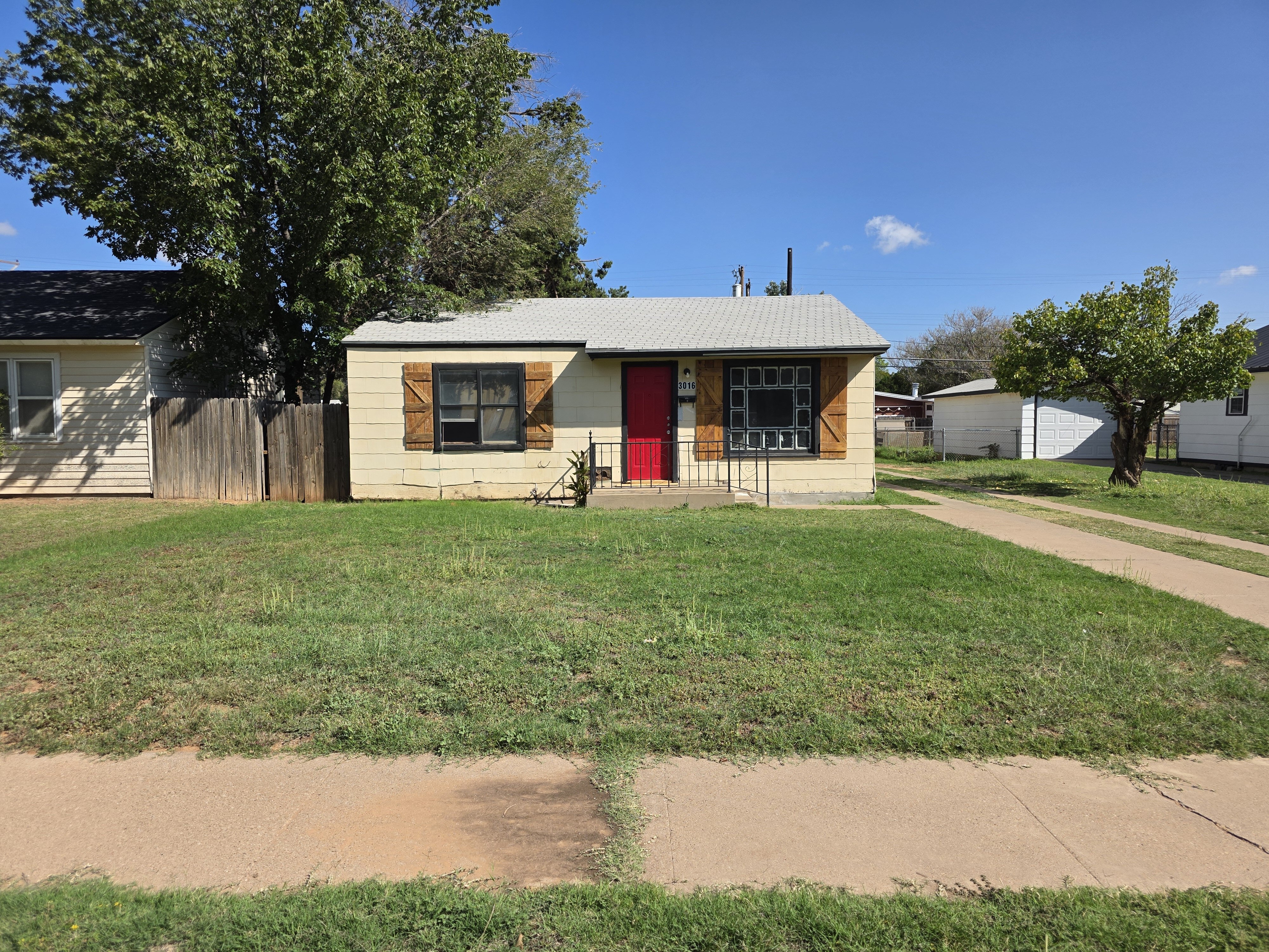 A small single-story yellow house in Lubbock, Texas with a bright red front door, wooden shutters, and a large front window. The home sits on a grassy lot with a long driveway extending to the backyard. A tall tree shades part of the house, and neighboring homes are visible on both sides under a clear blue sky.