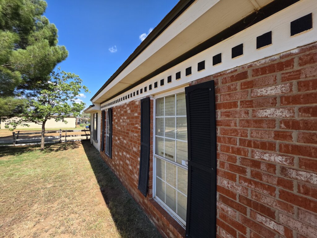 A close-up view of a red brick house in Lubbock, Texas, showing large windows with black shutters and decorative trim under the roofline. The side yard features a tree, a wooden fence, and a bright blue sky in the background.