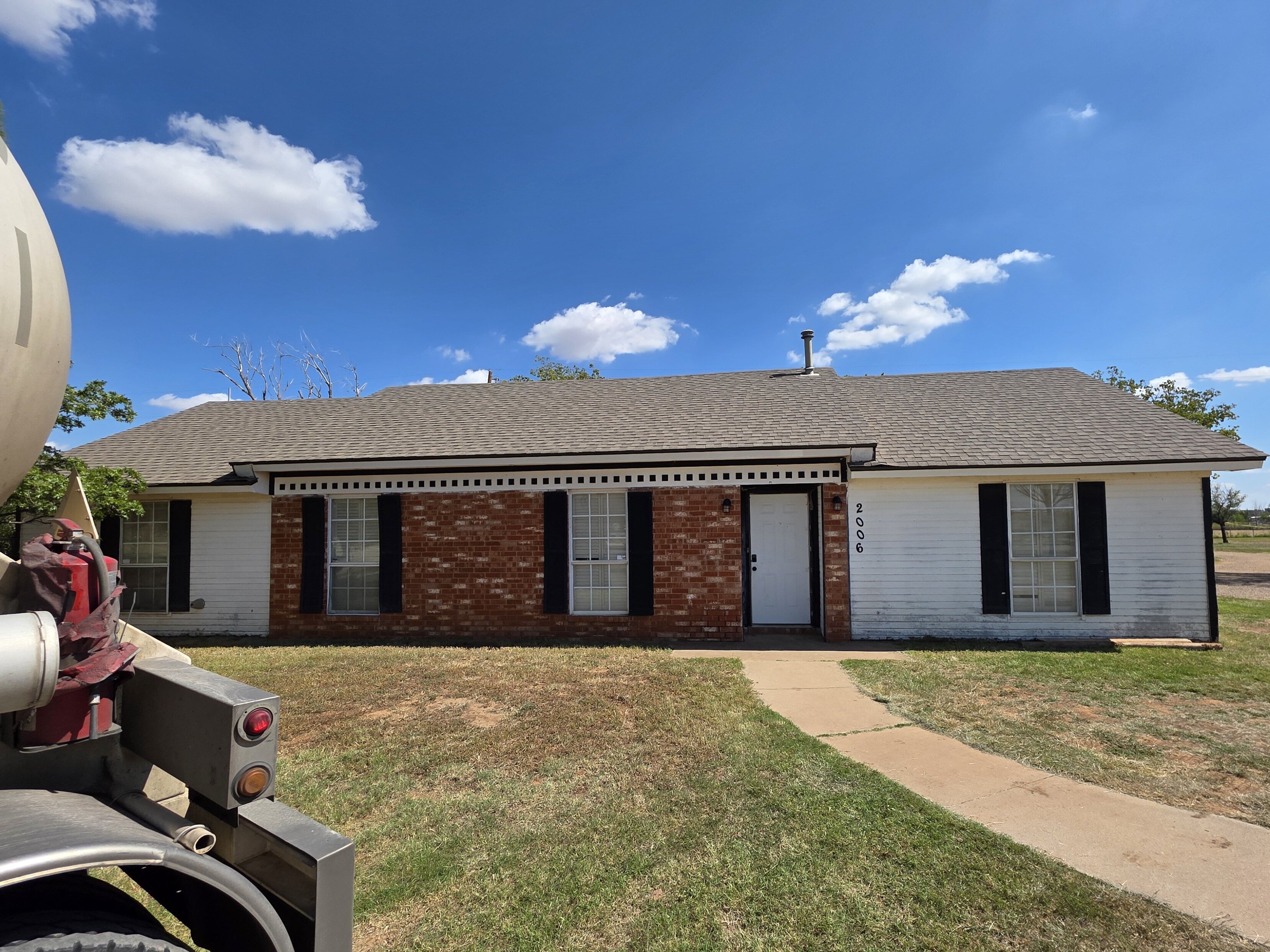 A single-story brick and white-siding house with black shutters under a bright blue sky in Lubbock, Texas. A curved concrete walkway leads to the front door, and a large utility truck is partially visible on the left side of the frame. The yard is dry with patches of grass, and open land with trees sits in the background.