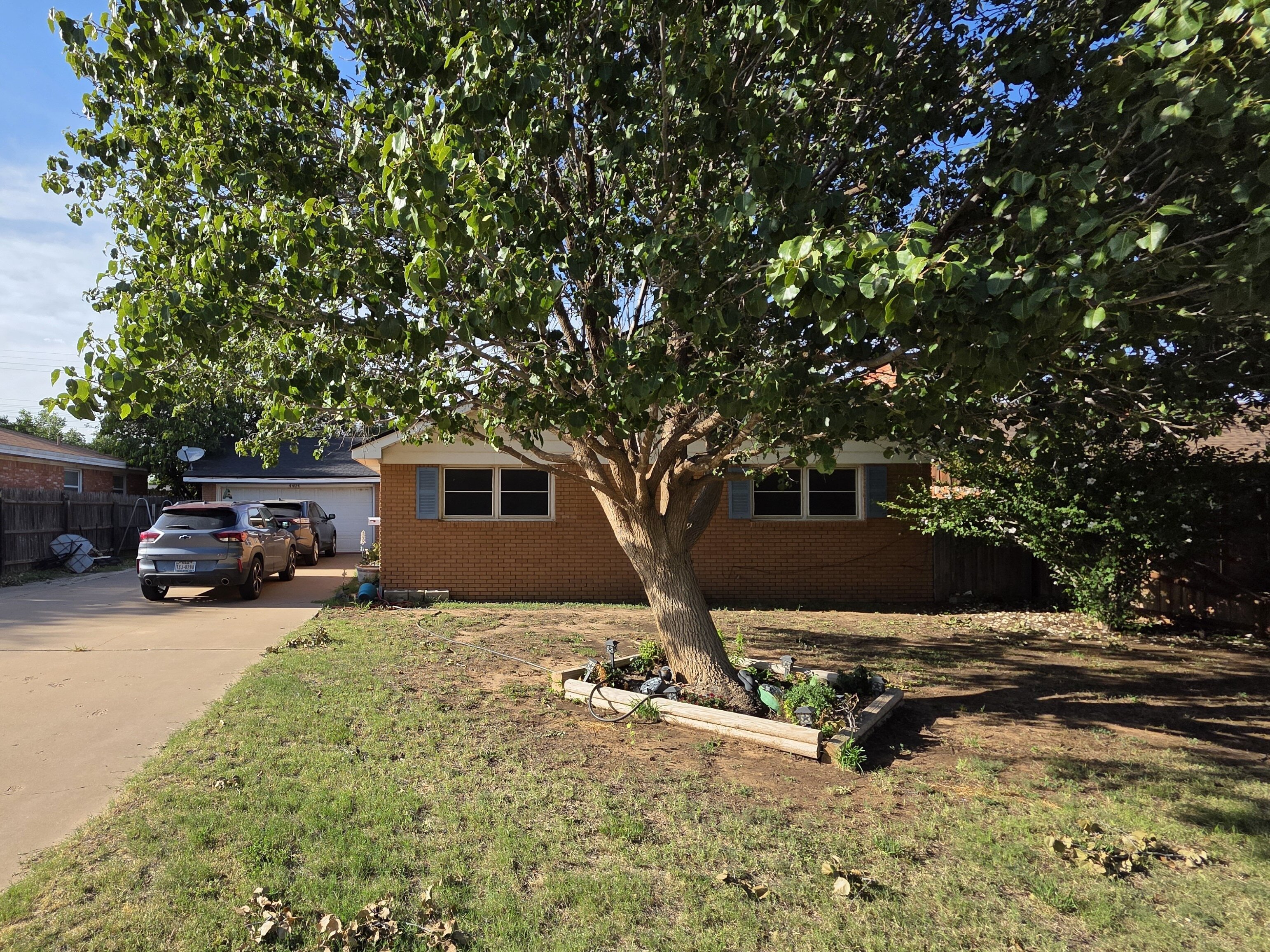 A single-story brick house in Lubbock, Texas with a large tree in the front yard casting shade over the lawn. Several vehicles are parked along the long driveway leading to the garage, and neighboring homes and fences are visible on each side under a bright blue sky.