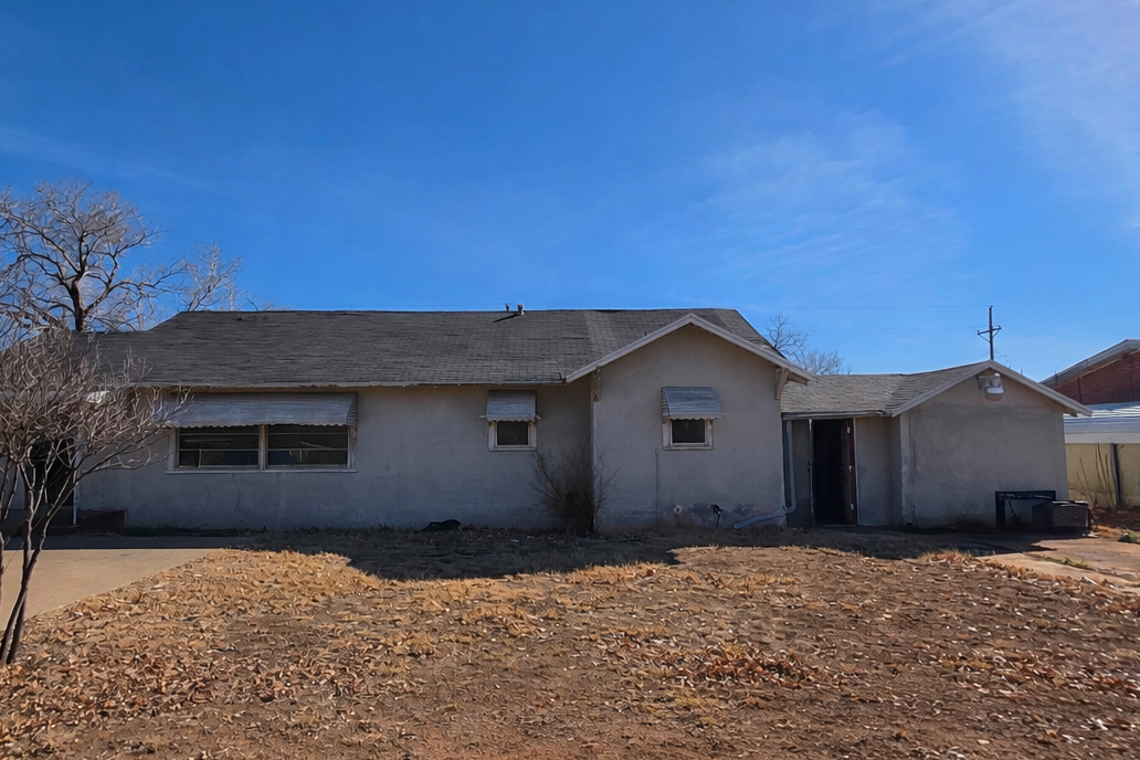 Front exterior of a single-story West Texas investment property in as-is condition with stucco siding, dry yard, and detached carport under a clear blue sky