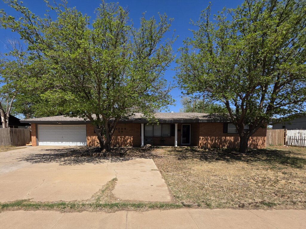 Single-story brick home in Lubbock, Texas, with an attached garage, mature trees, and a wide driveway.