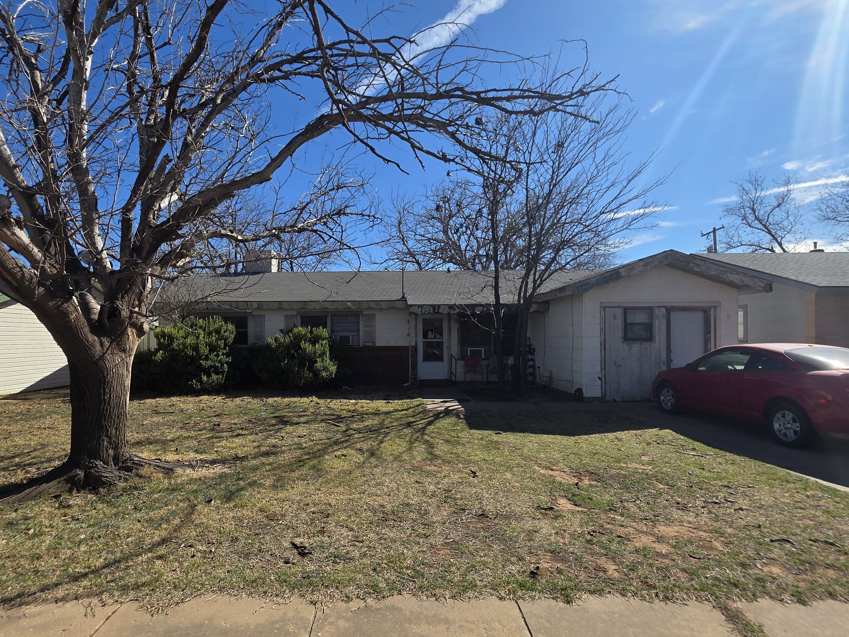 Older single-story home in Lubbock, Texas, with an attached garage, large front tree, and driveway parking.