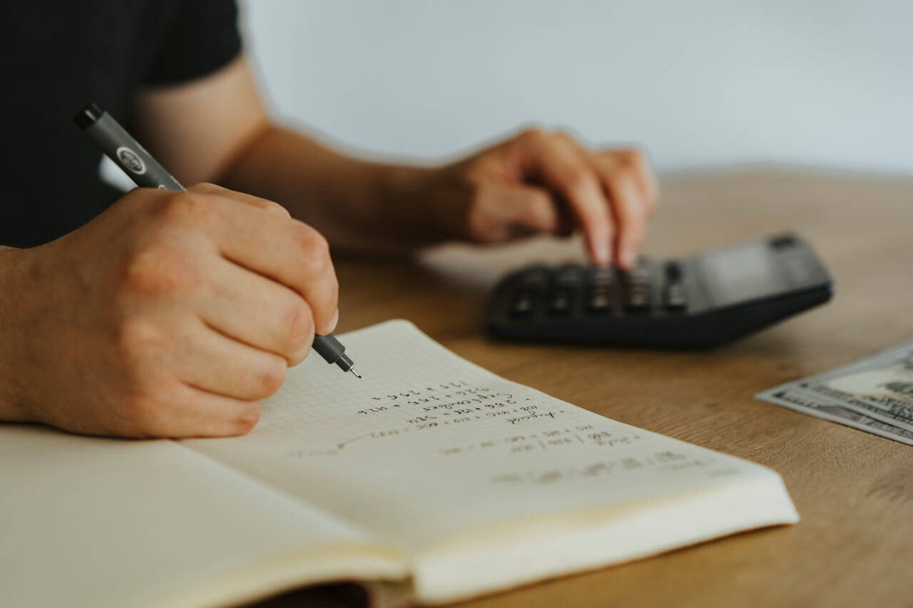 A person seated at a desk, writing in a journal and doing math on a calculator.