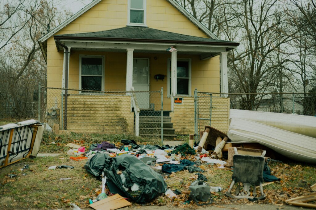 A distressed home with yellow siding. Trash and discarded furniture litter the front lawn.