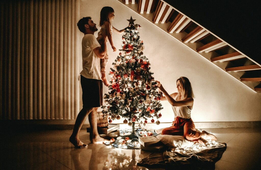 A father lifting his small daughter to place the start atop a festive Christmas tree. Nearby, a mother kneels to place ornaments on the other side of the tree.