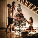 A father lifting his small daughter to place the start atop a festive Christmas tree. Nearby, a mother kneels to place ornaments on the other side of the tree.