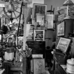 An inside look at a real hoarder's home, shown in black and white. Inside are stacks of framed pictures, appliances, books, dishes, and more. Possessions are piled dangerously high, with only a narrow walkway between.