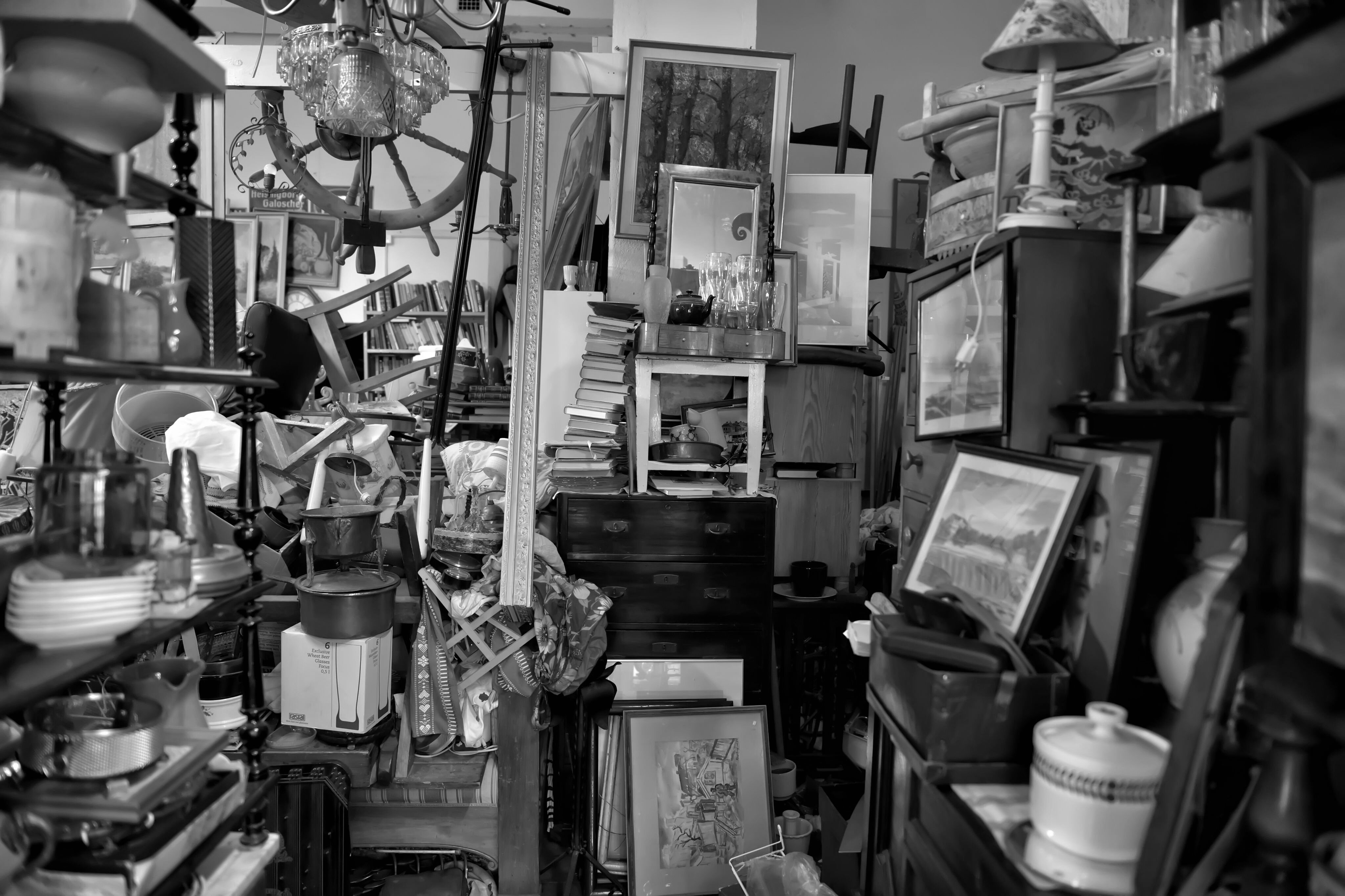 An inside look at a real hoarder's home, shown in black and white. Inside are stacks of framed pictures, appliances, books, dishes, and more. Possessions are piled dangerously high, with only a narrow walkway between.