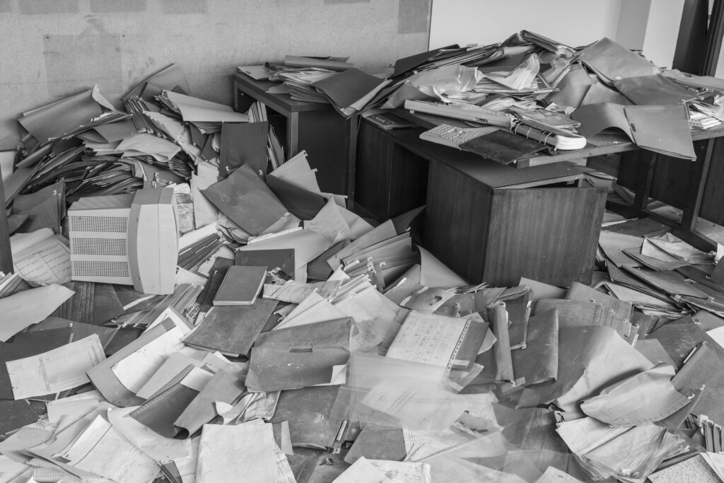 A real image from inside a hoarder's home, shown in black and white. An office with a desk and several tables, all piled high with loose paper, files, binders, and computer equipment, including monitors and keyboards. There is no clear pathway through the room shown.