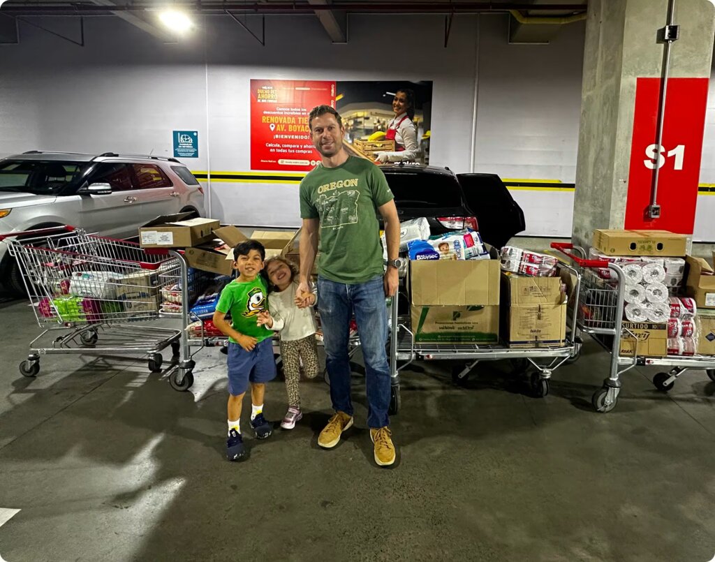 Scott Dalinger wearing an Oregon t-shirt stands smiling in a parking garage holding hands with his two young children, surrounded by shopping carts overflowing with donated relief supplies.