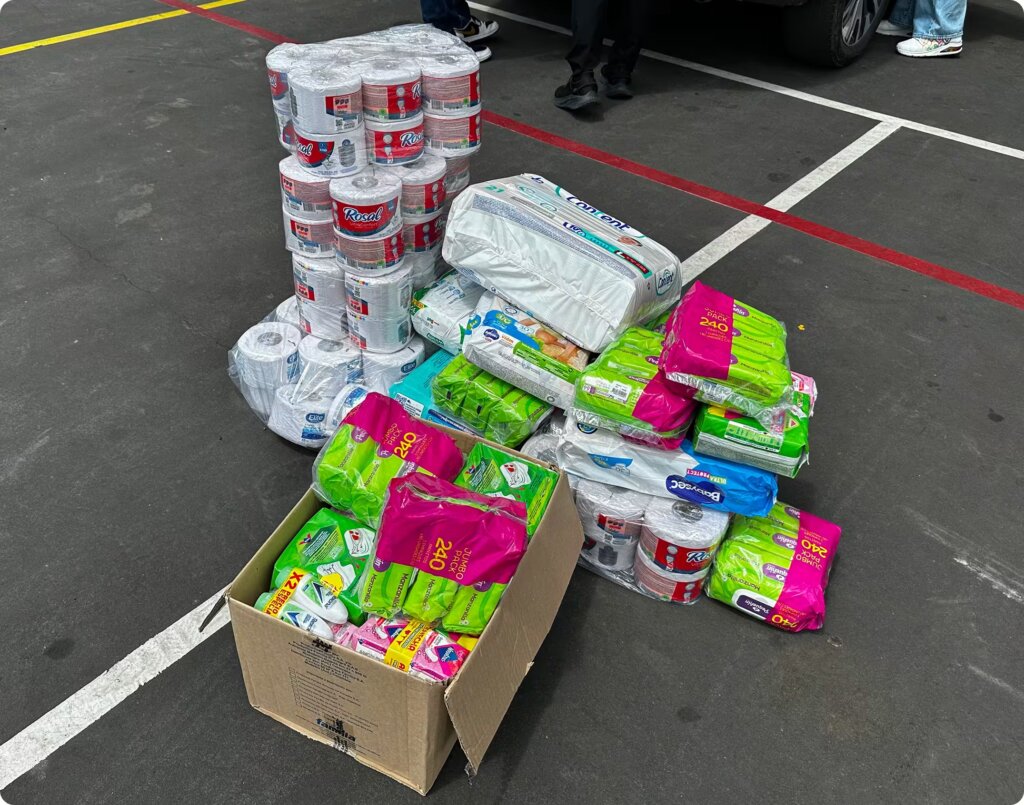 A large pile of essential supplies placed on asphalt, including stacks of toilet paper rolls, packs of baby diapers, and open boxes filled with feminine hygiene products.