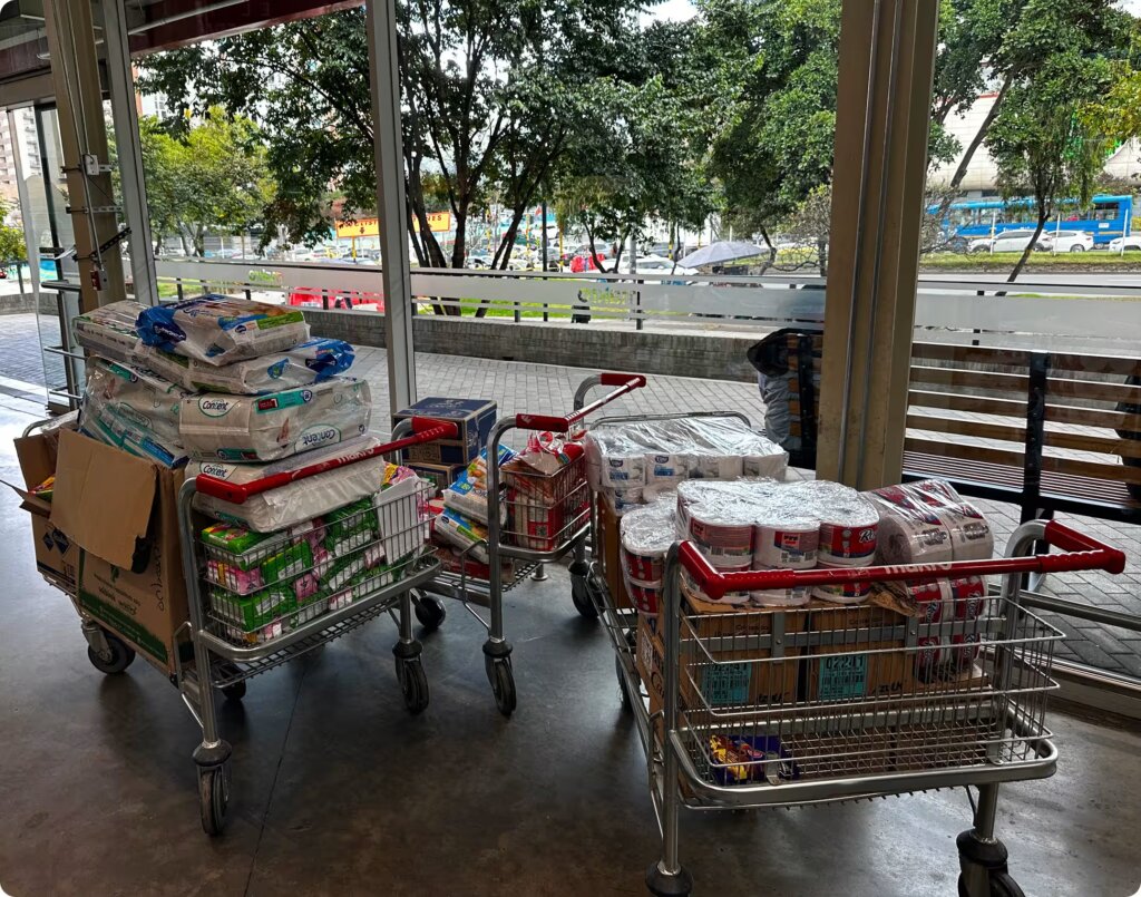 Three shopping carts lined up near a large glass window, stacked high with bulk packages of paper towels, toilet paper, adult diapers, and boxed food items.