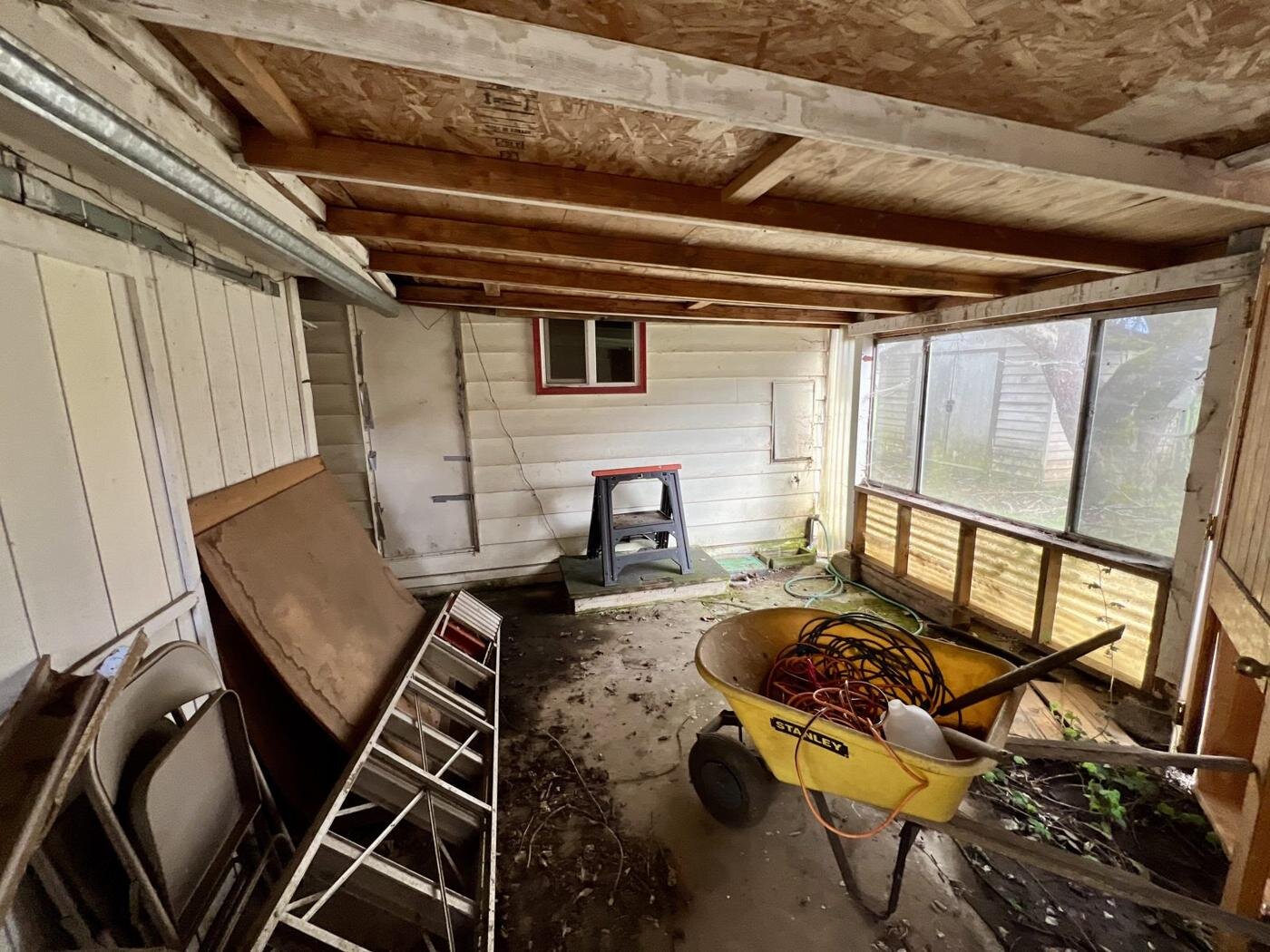Covered back porch used for storage, with a wheelbarrow and ladders