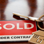 In this image, a key sits atop a glossy wooden surface, attached to a red tag boldly declaring "SOLD UNDER CONTRACT," symbolizing the successful sale of a property and the transition to new ownership.