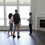 In the photo, a couple is attentively listening to a real estate agent who's gesturing and providing information about a modern, well-lit room.