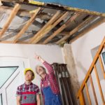 The image shows two construction workers inside a house, examining the exposed ceiling structure.