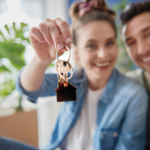 A happy couple is seen in the background, smiling and holding up a set of keys with a house-shaped keychain, symbolizing their excitement and joy as homeowners.