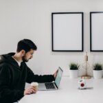 a man in a black hoodie sitted in a black chair ,holding a coffee cup while looking at computer on a white table.How to Sell My Land Yourself In Huntsville Alabama - We Buy Houses In Bama