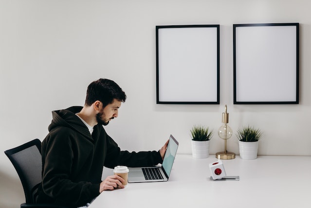 a man in a black hoodie sitted in a black chair ,holding a coffee cup while looking at computer on a white table.How to Sell My Land Yourself In Huntsville Alabama - We Buy Houses In Bama