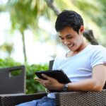 man in a white shirt on tablet smiling while sitting on black bistro outdoor chair with trees in the background. 5 Creative Ways To Sell Your Land Online