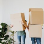 two people carrying cardboard boxes with plants in the left with white walls in the background. The lady has one box and a green t-shirt while the man has two boxes.5 Things You Can Do With The Proceeds From Your Vacant Land Sale In Huntsville