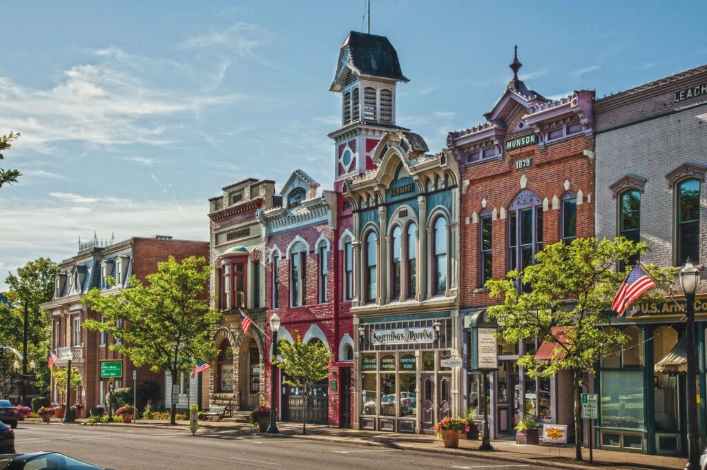 This is the header photo on Realty Done by Damien Baden's webpage for Luxury Homes for Sale in Medina, Ohio. It features a picturesque summertime streetscape in downtown Medina, Ohio, featuring well-preserved historic buildings with ornate architectural details. Located in Public Square along E Washington Street, the buildings are engraved with names and dates, such as 'Gazette 1835-1895,' 'Munson 1879' and 'Reinhardt 1888.' Businesses occupying these buildings include a dog bakery, gourmet popcorn shop, and the Town Hall & Engine House Museum. The sidewalk is lined with old-fashioned lamp posts, American flags, trees and planters with colorful flowers. The photo perfectly captures the beauty and small-town America charm of downtown Medina.