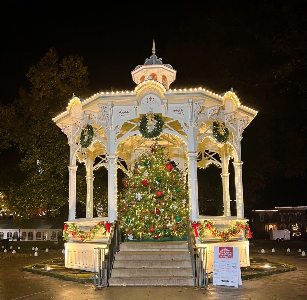This is a photo on Realty Done by Damien Baden's webpage for Luxury Homes for Sale in Medina, Ohio. It features the iconic white gazebo in historic downtown Medina's Uptown Park all decorated for Christmas. The gazebo is adorned with garland, wreaths, white lights, and a large Christmas tree with red, green, and gold ornaments stands inside. A sign at the bottom right of the steps lists sponsors for the holiday display. The scene is illuminated against a dark night sky, creating a festive and inviting atmosphere.