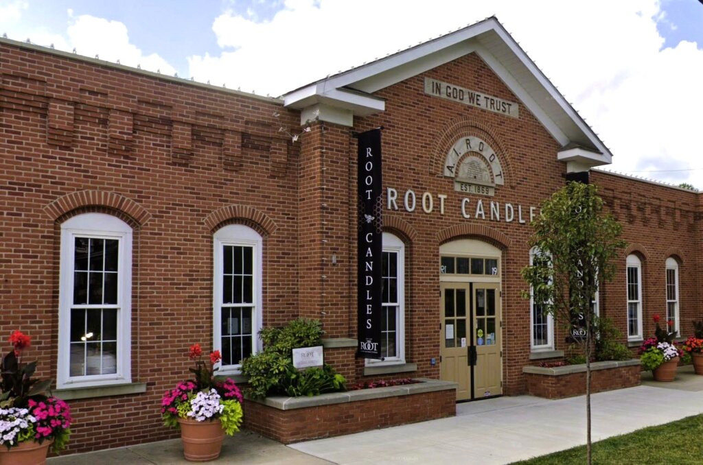 This is a photo on Realty Done by Damien Baden's webpage for Luxury Homes for Sale in Medina, Ohio. This image shows the exterior of Root Candles, a historic building in downtown Medina, Ohio. The building's architecture features a red brick facade with arched windows and a double-door entrance. Decorative planters with colorful flowers enhane its curb appeal. Dating back to 1869, the signage indicates the establishment's long history as Ohio's oldest candle manufacturer.