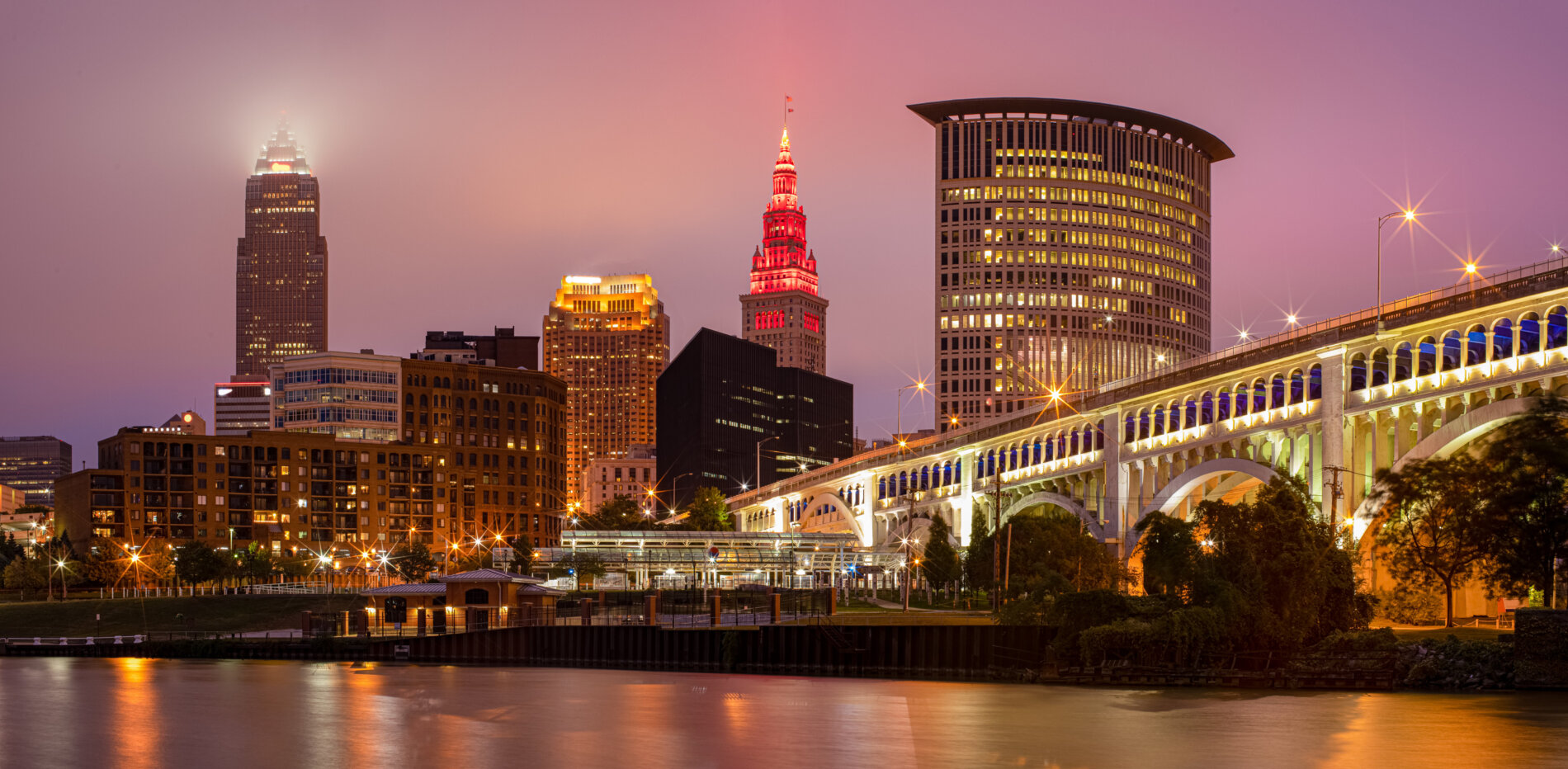 Downtown Cleveland skyline at dusk with illuminated skyscrapers and the brightly lit Veterans Memorial Bridge over The Cuyahoga River. This is the cover photo for Realty Done by Damien Baden's homepage – the best real estate agent to help you buy and sell homes in Northeast Ohio.