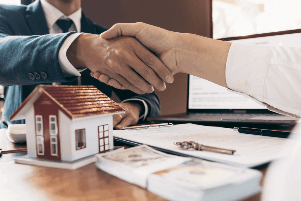Business handshake over a desk with a house model, contract papers, and money, symbolizing a real estate agreement