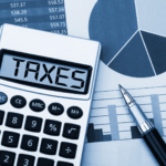 Calculator displaying the word taxes beside financial charts and a pen.