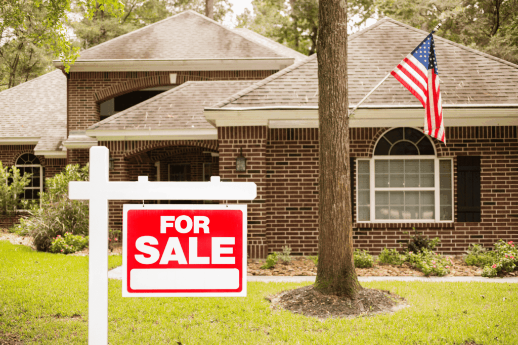 For sale sign in front of a brick house with an American flag.