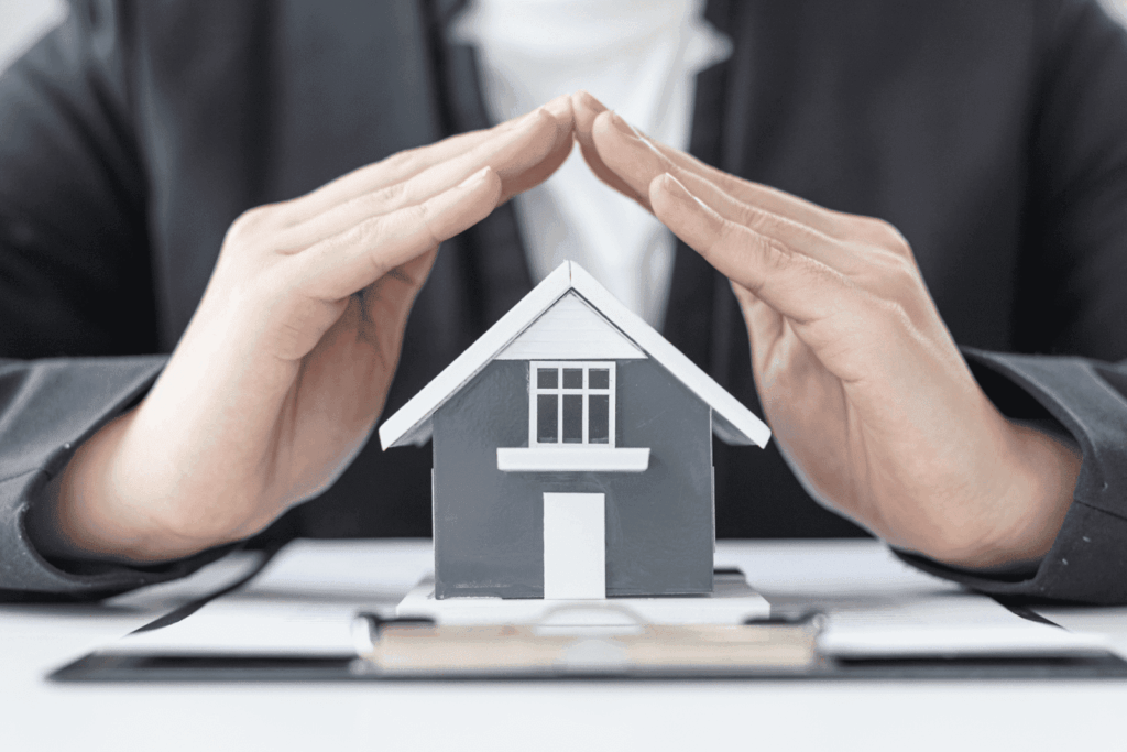 Hands forming a roof shape over a small house model symbolizing home protection.