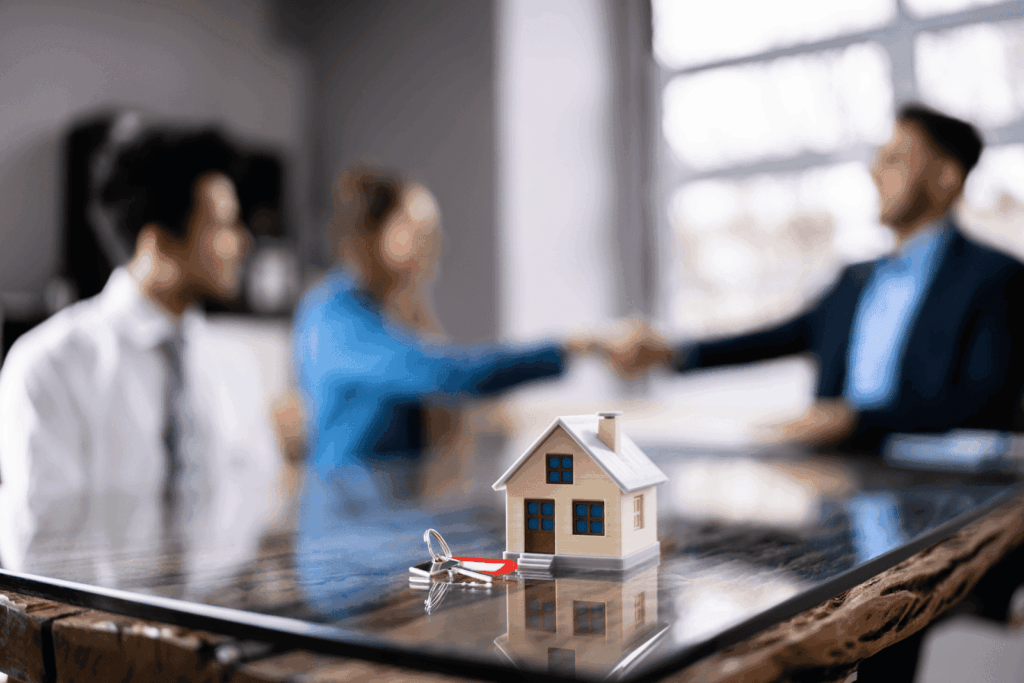 Mini house model and keys on table with people shaking hands in background