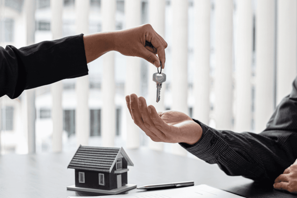 Person handing over house keys to another person with a small house model on the table.
